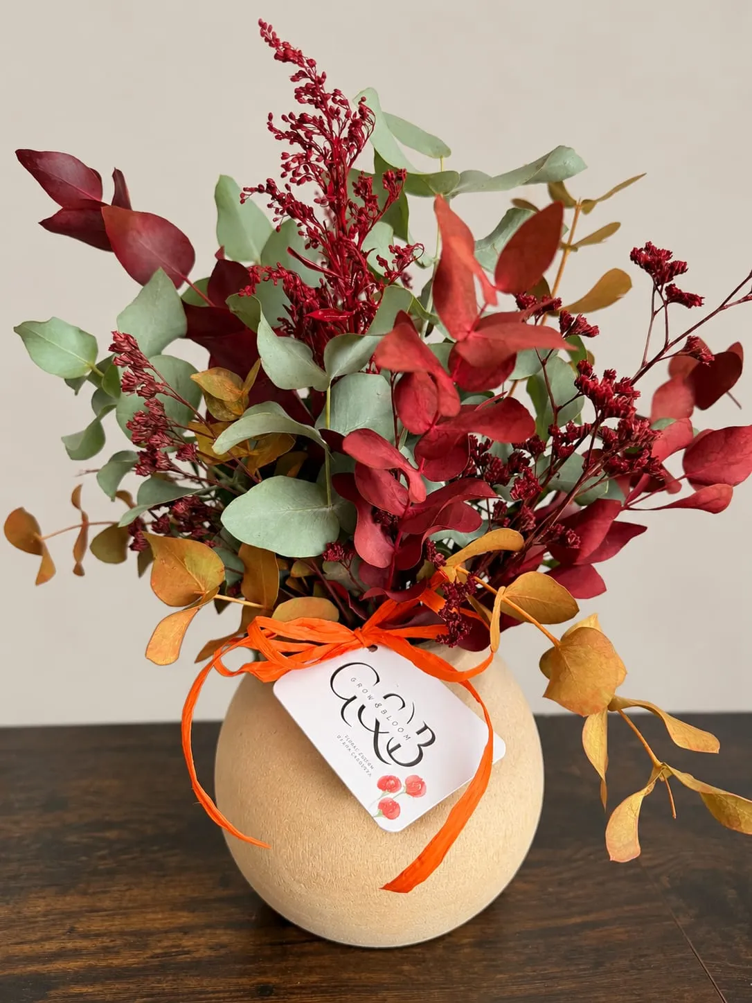 Jarrón redondo de cerámica beige con flores preservadas en rojo, granate, ocre y verde: eucalipto, statice y espigas teñidas, atado con rafia naranja y la tarjeta de Grow & Bloom