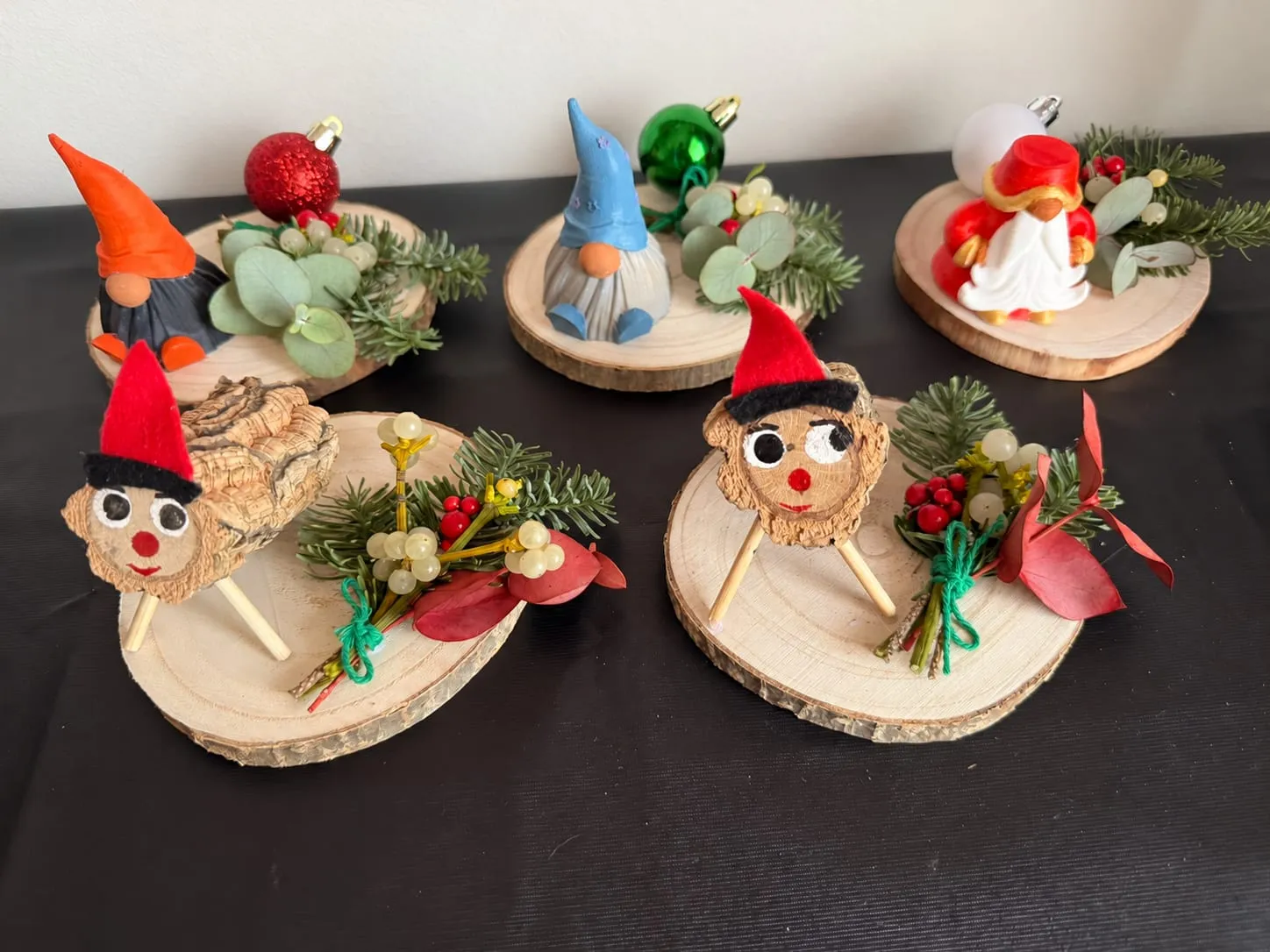 Cinco centros navideños sobre rodajas de tronco: dos tions de corcho con barretina roja y cara pintada, dos gnomos y un Papá Noel, decorados con abeto, eucalipto, muérdago, bayas rojas y bolas de Navidad