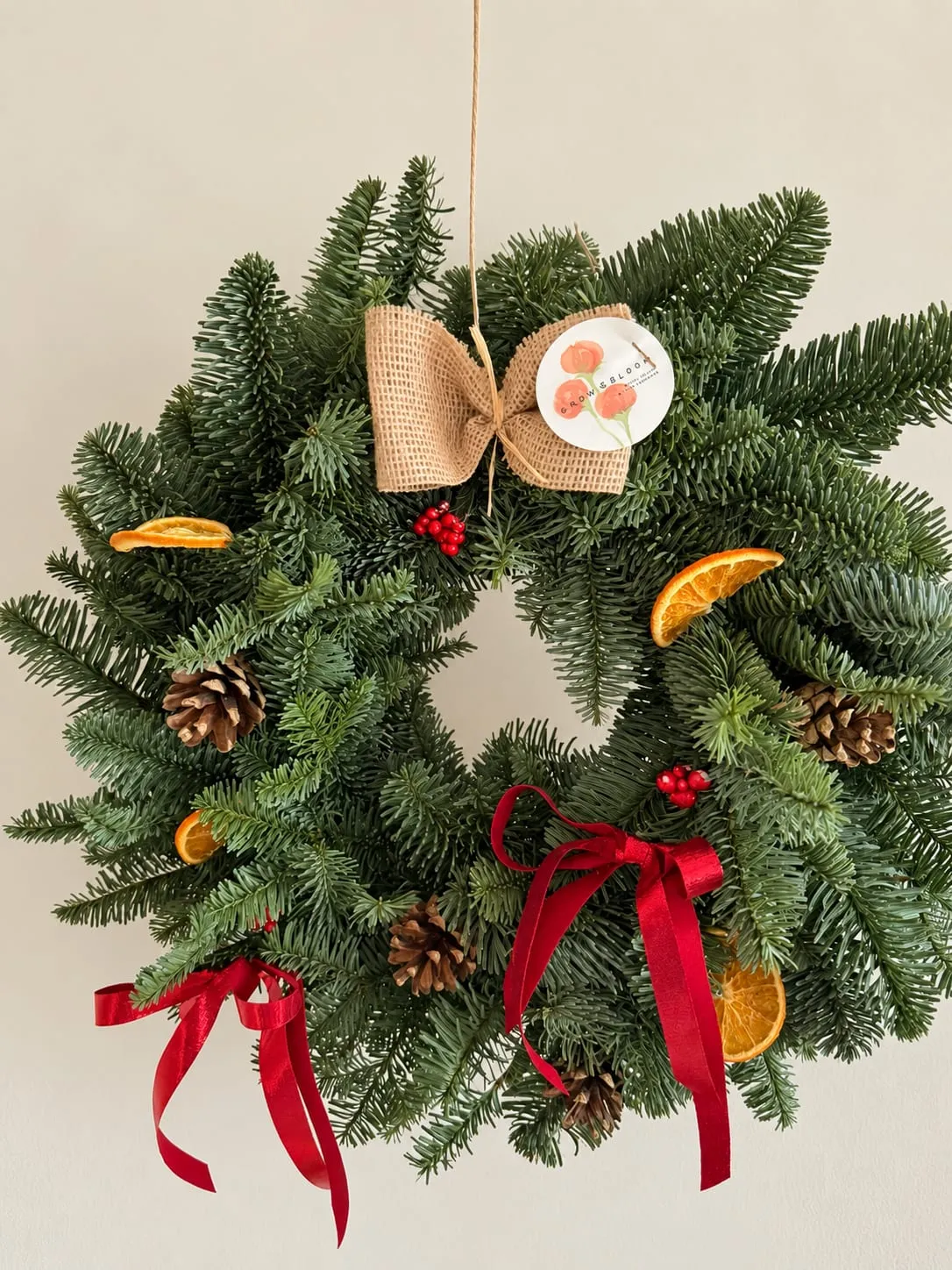 Corona de Navidad de abeto colgada en la pared, con rodajas de naranja seca, piñas, bayas rojas, lazos de raso rojo y un lazo de arpillera con la etiqueta de Grow & Bloom
