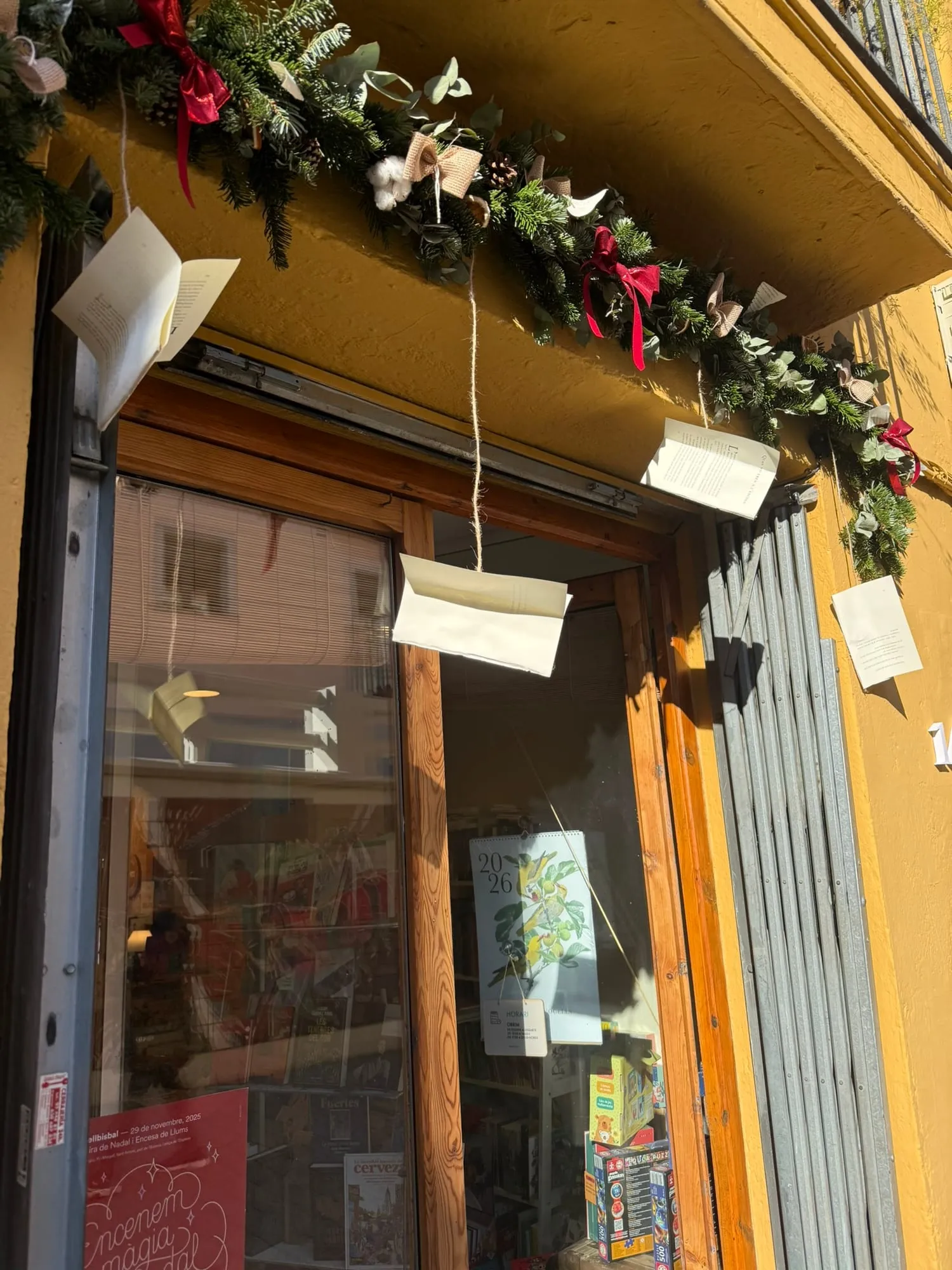Guirnalda navideña de abeto, eucalipto, piñas, algodón y lazos rojos y de arpillera sobre la puerta de una librería de fachada amarilla, con páginas de libro colgando de cordeles