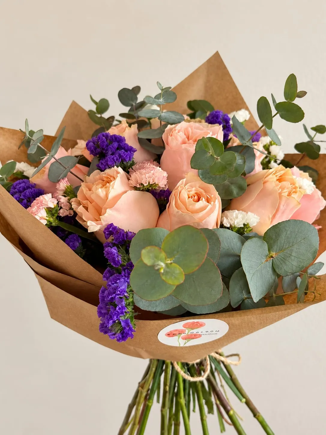 Ramo envuelto en papel kraft con rosas de jardín en tono melocotón, statice morado, clavelinas rosas y blancas y eucalipto, atado con cordel y con la pegatina de Grow & Bloom