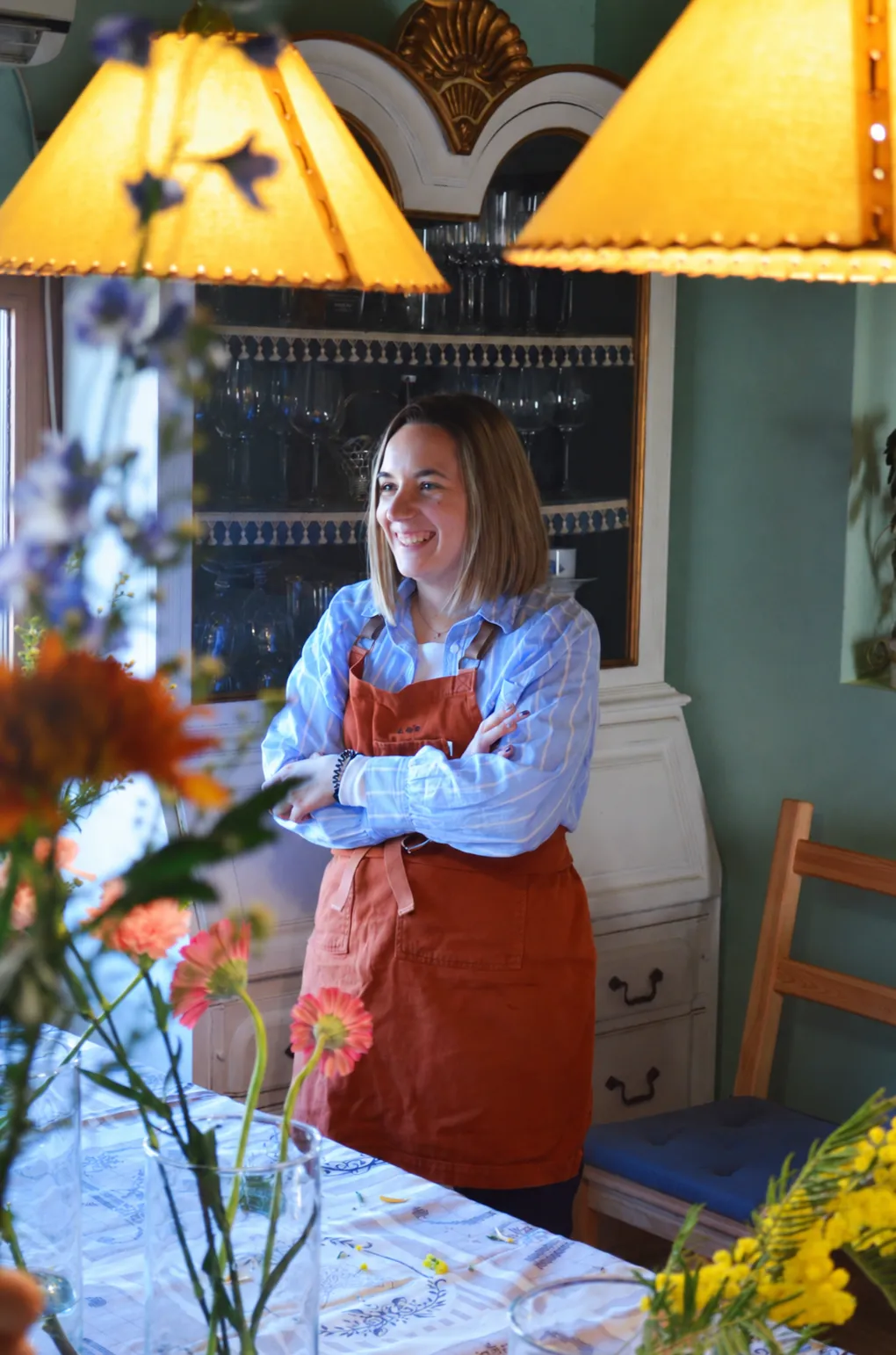 Laia sonríe de pie, con los brazos cruzados y un delantal teja, junto a una mesa con flores.
